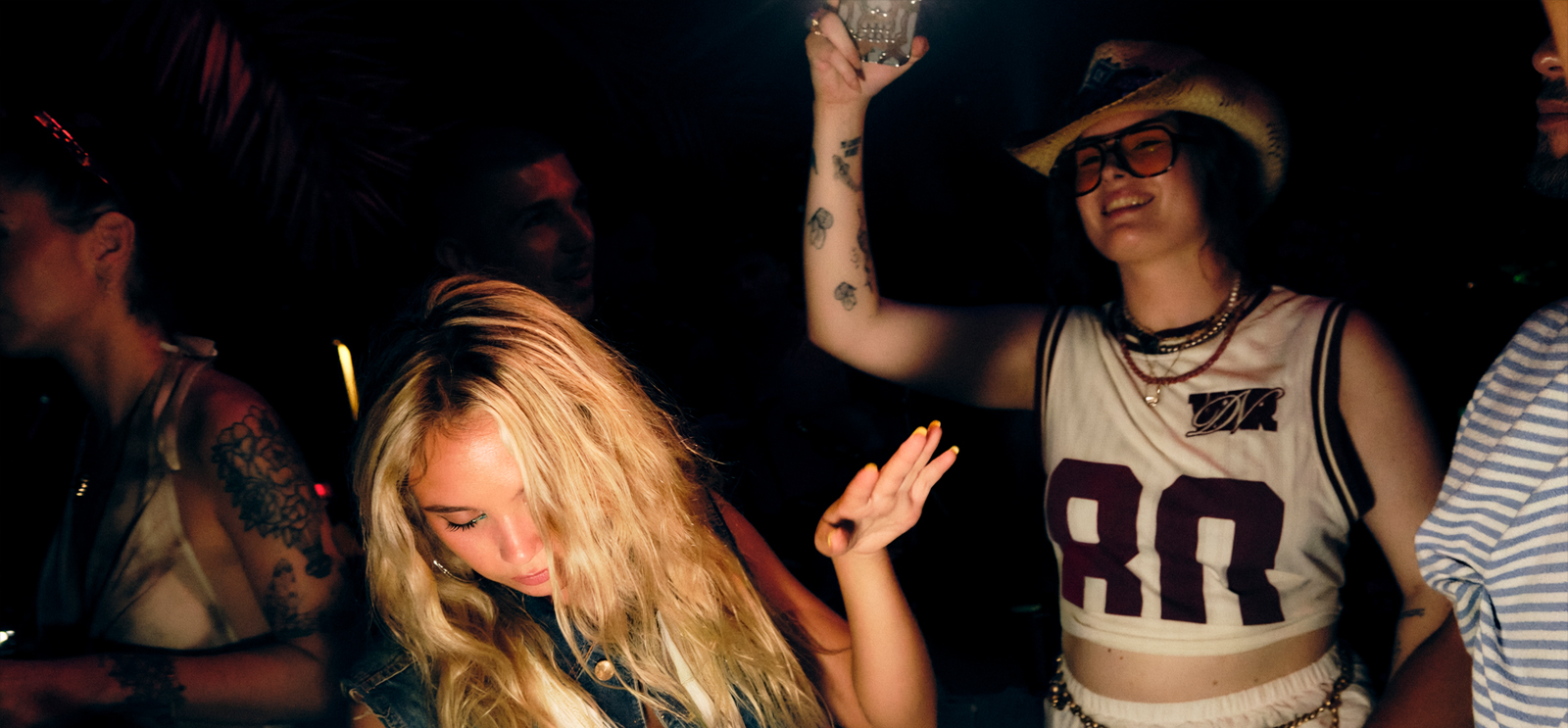 Pareja joven sonriendo en una fiesta nocturna; ella tapa su copa con la mano